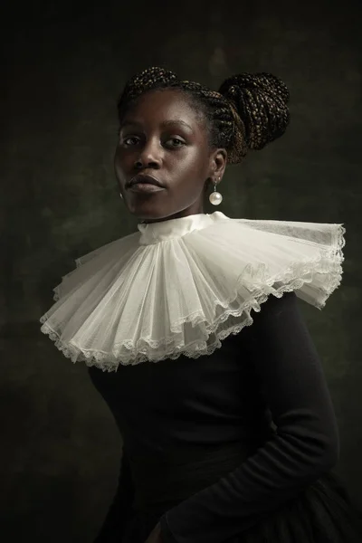 Portrait of medieval African young woman in black vintage dress with big white collar posing isolated on dark green background.