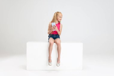 Beyaz stüdyo arka planında izole edilmiş bir kutunun üzerinde günlük kıyafetlerle oturan güzel küçük bir kız. Mutlu çocukluk kavramı.