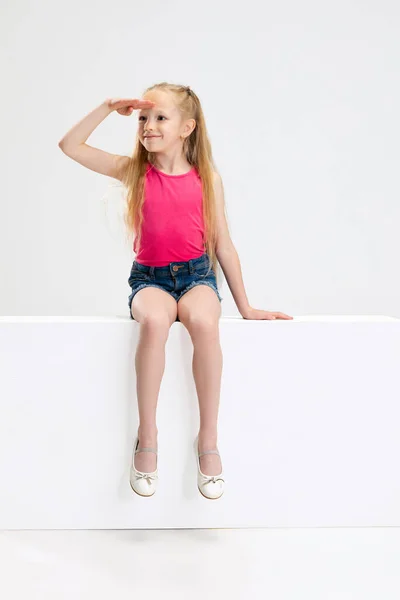 Une belle petite fille en tenue décontractée assise sur une boîte isolée sur fond de studio blanc. Concept d'enfance heureuse. — Photo