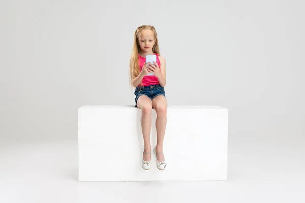 Jedna piękna dziewczynka w luźnych ubraniach siedzi na pudełku odizolowany na białym tle studio. Szczęśliwego dzieciństwa. — Zdjęcie stockowe