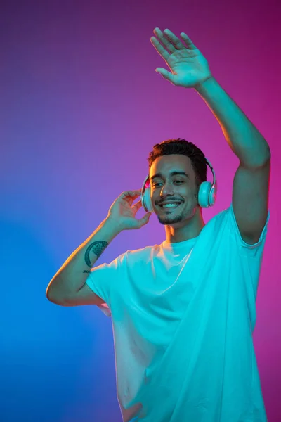 Retrato de jovem latino em fones de ouvido brancos posando isolado no gradiente roxo fundo rosa em luz de néon. — Fotografia de Stock