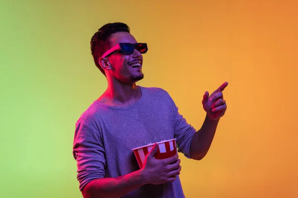 Portrait of Latina young man in 3d glasses holding bucket of popcorn isolated on gradient yellow green background in neon light. — Stock Photo, Image