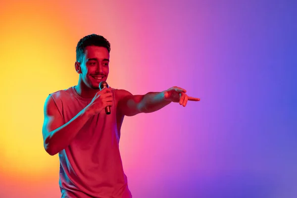 Portrait of Latina young man, singer with microphone performing isolated on gradient yellow purple background in neon light. Concept of emotions, facial expression — Stock Photo, Image