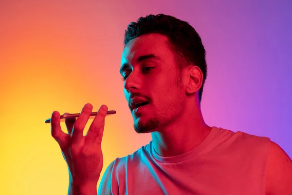 Porträtt av Latina ung man med telefon, smartphone isolerad på lutning gul lila bakgrund i neon ljus. Begreppet känslor, ansiktsuttryck — Stockfoto