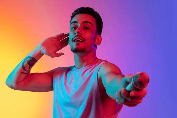 Retrato do jovem latino, cantor com microfone apresentando-se isolado em fundo púrpura amarelo gradiente em luz de néon. Conceito de emoções, expressão facial — Fotografia de Stock
