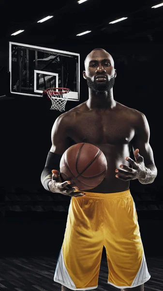 Porträt eines jungen afrikanischen Sportlers, Basketballspielers, der mit Ball in der Turnhalle stapft, Türen isoliert auf dunklem Hintergrund. Konzept von Sport, Spiel, Wettbewerb. — Stockfoto