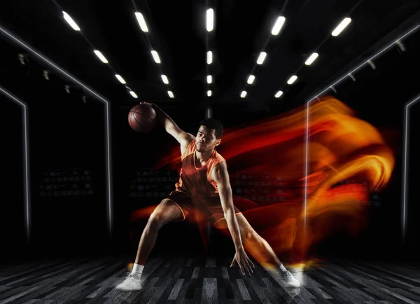 Jeden mladý sportovec basketbalista trénink v tělocvičně, idoors izolované na tmavém pozadí ve smíšeném světle. Pojetí sportu, hry, soutěže. — Stock fotografie