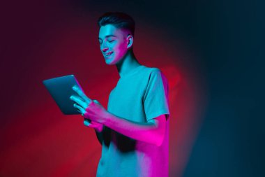 Tabletli beyaz bir gencin portresi. Cihaz, koyu kırmızı stüdyo arka planında, neon pembe ışıkta izole edilmiş. İnsan duyguları kavramı, yüz ifadesi.