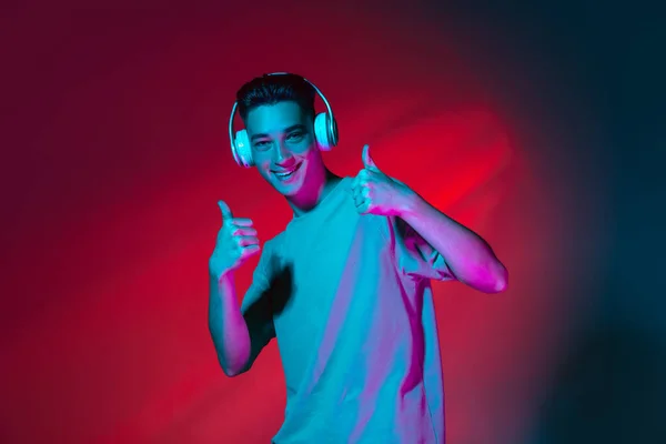 Retrato do jovem caucasiano em fones de ouvido isolados no fundo do estúdio vermelho escuro em luz rosa néon. Conceito de emoções humanas, expressão facial. — Fotografia de Stock