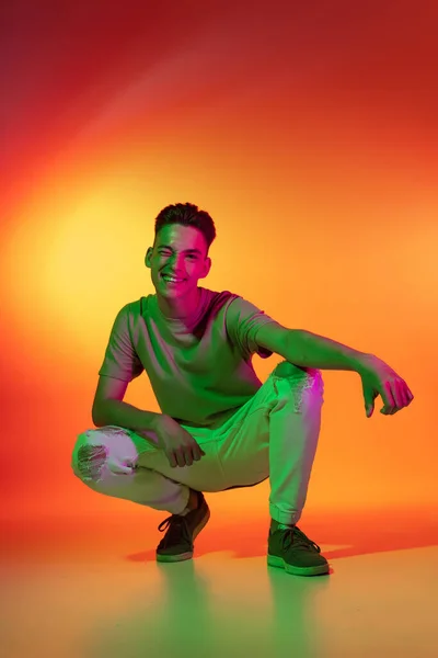 Retrato do jovem caucasiano sentado isolado no fundo do estúdio laranja em luz de néon. Conceito de emoções humanas, expressão facial. — Fotografia de Stock