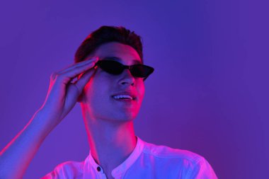 Güneş gözlüklü, beyaz tenli genç adam mavi stüdyo arka planında, neon pembe ışıkta izole edilmiş. İnsan duyguları kavramı, yüz ifadesi.