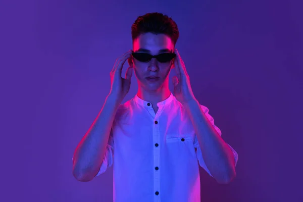 Biały młodzieniec w okularach przeciwsłonecznych odizolowany na niebieskim tle studia w neonowym, różowym świetle. Pojęcie ludzkich emocji, wyraz twarzy. — Zdjęcie stockowe