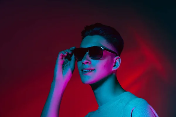 Porträt eines kaukasischen jungen Mannes mit Sonnenbrille isoliert auf dunkelrotem Studiohintergrund in neonrosa Licht. Konzept menschlicher Emotionen, Mimik. — Stockfoto