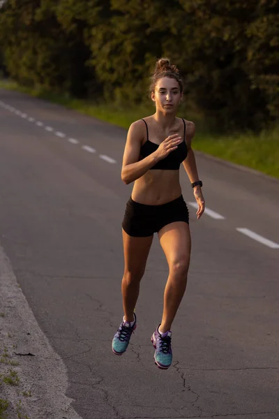 Young female runner, athlete is jogging at road in summer sunshine. Beautiful caucasian woman training, listening to music. Concept of sport, healthy lifestyle, movement, activity. — Stock Photo, Image