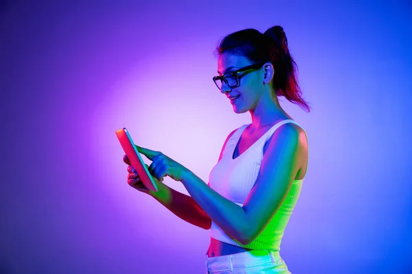Memegang tablet, gila bersemangat. Wanita muda kaukasia potret di latar belakang studio hitam dalam cahaya neon. Cantik model pria dengan kemeja putih. — Stok Foto