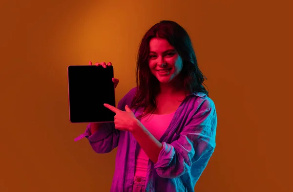 Holding tablet, crazy excited. Caucasian young womans portrait on black studio background in neon light. Beautiful male model in white shirt. — Stock Photo, Image