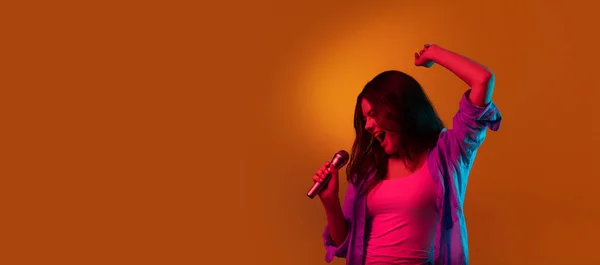 Portrait de chanteuse caucasienne isolé sur fond de studio orange au néon. Beau modèle féminin dans un style décontracté avec microphone. Concept d'émotions humaines — Photo