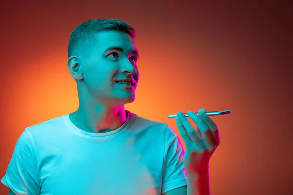 Nahaufnahme Porträt eines jungen kaukasischen Mannes mit Telefon isoliert über Gradienten rot-orangefarbenen Studiohintergrund in Neonlicht mit Kopierfläche für Werbung. Konzept menschlicher Emotionen — Stockfoto