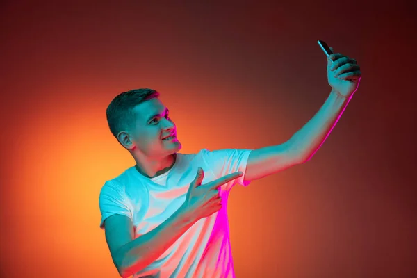 Retrato de jovem caucasiano usando telefone isolado sobre gradiente vermelho laranja estúdio fundo em luz de néon com copyspace para anúncio. Conceito de emoções humanas — Fotografia de Stock