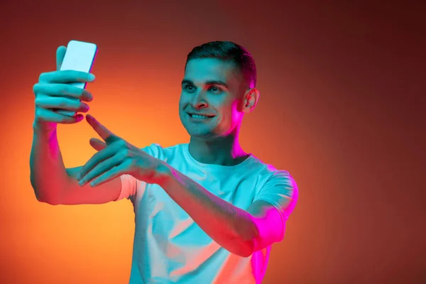 Retrato de jovem caucasiano usando telefone isolado sobre gradiente vermelho laranja estúdio fundo em luz de néon com copyspace para anúncio. Conceito de emoções humanas — Fotografia de Stock