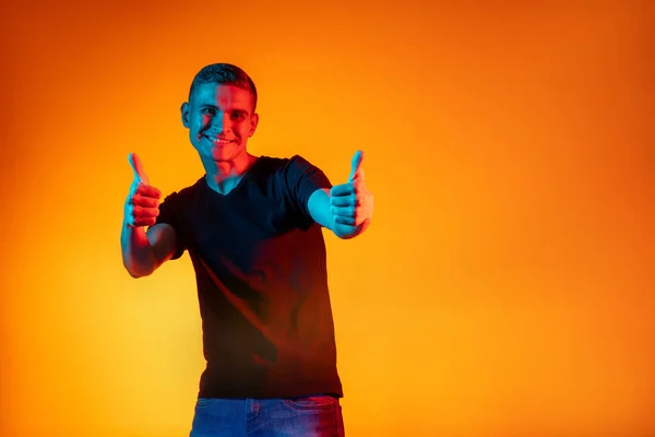 Retrato de un joven hombre caucásico en ropa casual aislado sobre fondo de estudio de color rojo anaranjado degradado en luz de neón con espacio para publicidad. Concepto de emociones humanas —  Fotos de Stock