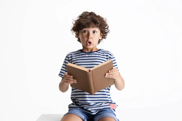 Un niño preescolar caucásico sentado y leyendo un libro aislado sobre fondo blanco del estudio. Copyspace. Infancia, educación, concepto de emociones — Foto de Stock