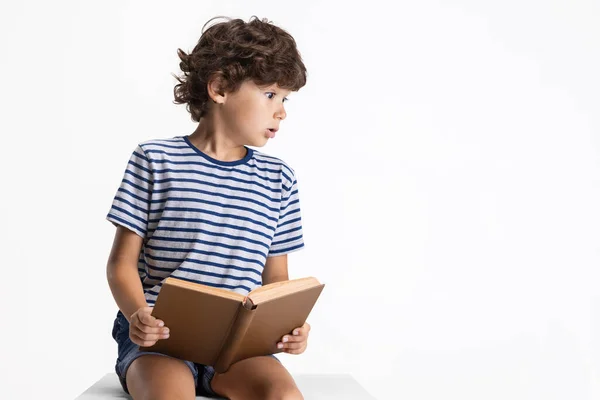 Un niño preescolar caucásico sentado y leyendo un libro aislado sobre fondo blanco del estudio. Copyspace. Infancia, educación, concepto de emociones —  Fotos de Stock