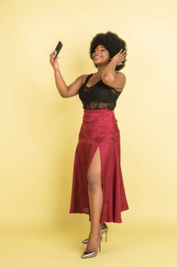 Açık sarı stüdyo arka planında izole edilmiş telefonuyla poz veren güzel Afrikalı genç bir kadın. İnsan duyguları kavramı, yüz ifadesi, gençlik, satış, reklam.