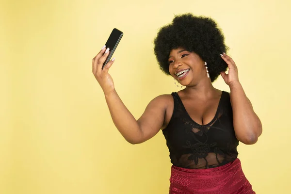 Hermosa mujer joven africana posando con teléfono aislado sobre fondo de estudio amarillo claro. Concepto de emociones humanas, expresión facial, juventud, ventas, anuncio. —  Fotos de Stock