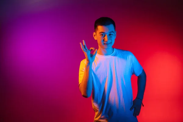 Un jeune homme asiatique isolé sur fond studio dans un dégradé de lumière rose-violet néon, filtre de couleur. Concept d'émotions humaines, expression faciale. — Photo