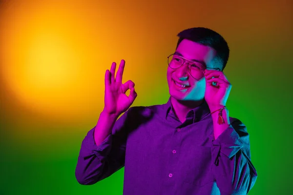 Portrait de jeune homme asiatique isolé en studio dans un dégradé de lumière jaune néon vert, filtre de couleur. Concept d'émotions humaines, expression faciale. — Photo
