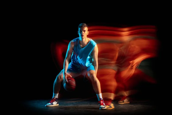 Junge kaukasische männliche Basketballspieler beim Basketballspielen isoliert vor dunklem Studiohintergrund in gemischtem Licht. Konzept von gesundem Lebensstil, Profisport, Hobby. — Stockfoto