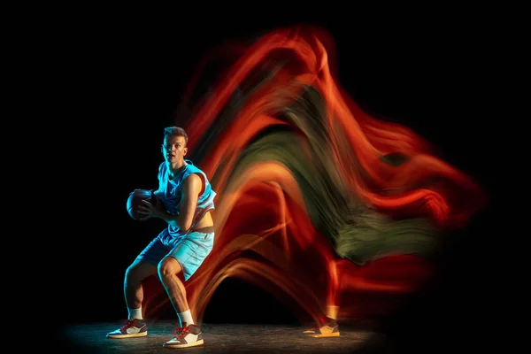 Professional male basketball player in blue uniform playing basketball isolated over dark studio background in mixed light. Concept of healthy lifestyle, professional sport, hobby.