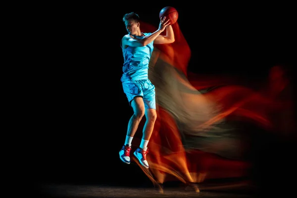 Mladý kavkazský hráč basketbalu hraje basketbal izolovaný přes tmavé pozadí studia ve smíšeném světle. Koncept zdravého životního stylu, profesionální sport, hobby. — Stock fotografie