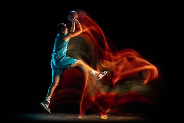 Junge kaukasische männliche Basketballspieler beim Basketballspielen isoliert vor dunklem Studiohintergrund in gemischtem Licht. Konzept von gesundem Lebensstil, Profisport, Hobby. — Stockfoto