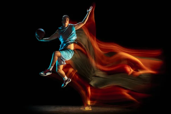 Jeune joueur caucasien de basket-ball masculin jouant au basket isolé sur fond de studio sombre dans une lumière mélangée. Concept de mode de vie sain, sport professionnel, passe-temps. — Photo