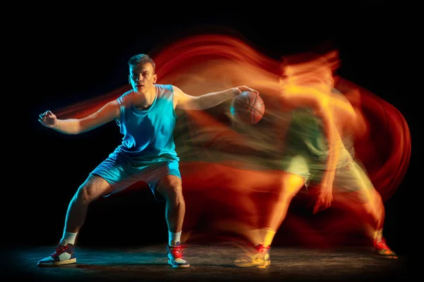 Young caucasian male basketball player playing basketball isolated over dark studio background in mixed light. Concept of healthy lifestyle, professional sport, hobby. Stock Photo