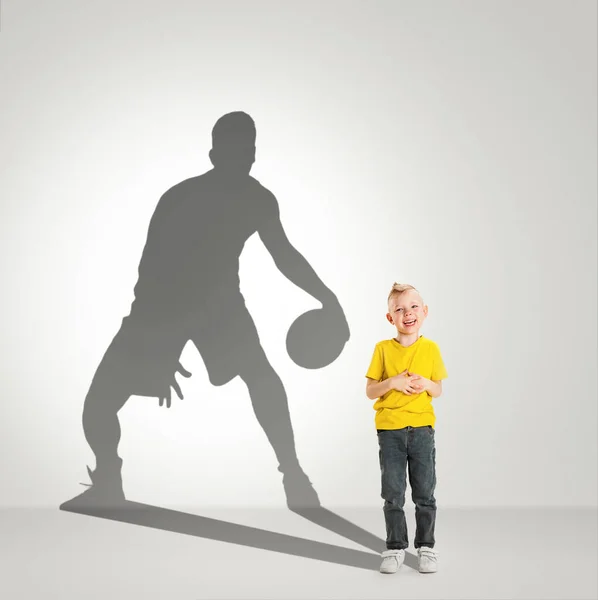 Мечтает о большом и известном будущем. Концептуальное изображение с маленьким мальчиком и тенью подходящего баскетболиста на светло-сером фоне — стоковое фото