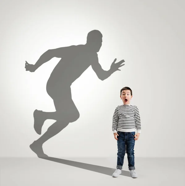 Childhood and dream about big and famous future. Conceptual image with boy and shadow of fit male runner on light gray background — Stockfoto