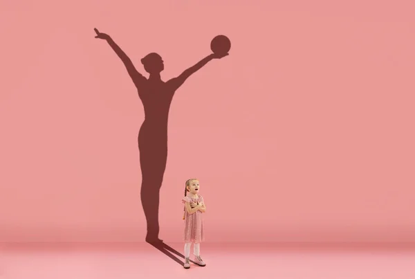Παιδική ηλικία και όνειρο για μεγάλο και διάσημο μέλλον. Εννοιολογική εικόνα με το κορίτσι και τη σκιά της ταιριάζει θηλυκό γυμναστή ρυθμό σε κοραλί ροζ φόντο — Φωτογραφία Αρχείου