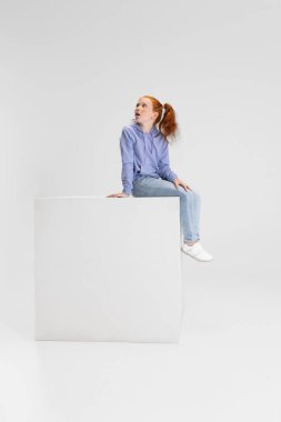Beyaz stüdyo arka planında izole edilmiş büyük bir kutunun üzerinde oturan, gündelik kıyafetli, tatlı kızıl saçlı bir kız. Mutlu çocukluk kavramı. Neşeli çocuk. Mutlu görünüyor, mutlu.