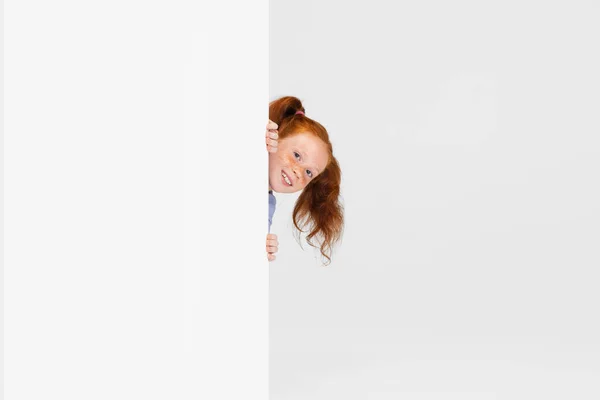Porträtt av söt fräknig rödhårig flicka i casual outfit kikar ut väggen isolerad på vit studio bakgrund. Glad barndomsidé. Soligt barn — Stockfoto
