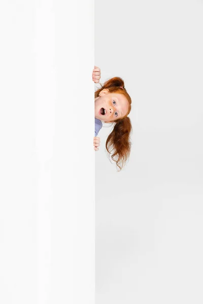 Portret van schattig sproeten roodharige meisje in casual outfit gluuren uit de muur geïsoleerd op witte studio achtergrond. Gelukkige kindertijd concept. Zonnig kind — Stockfoto