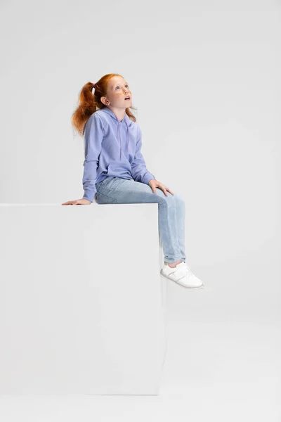 Uma linda menina ruiva em roupas casuais sentada na caixa grande isolada no fundo do estúdio branco. Feliz conceito de infância. Criança ensolarada. Parece feliz, encantado — Fotografia de Stock