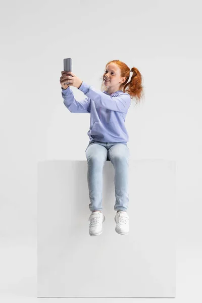 One cute red-headed girl in casual clothes sitting on big box isolated on white studio background. Happy childhood concept. Sunny child. Looks happy, delighted — Stock Photo, Image