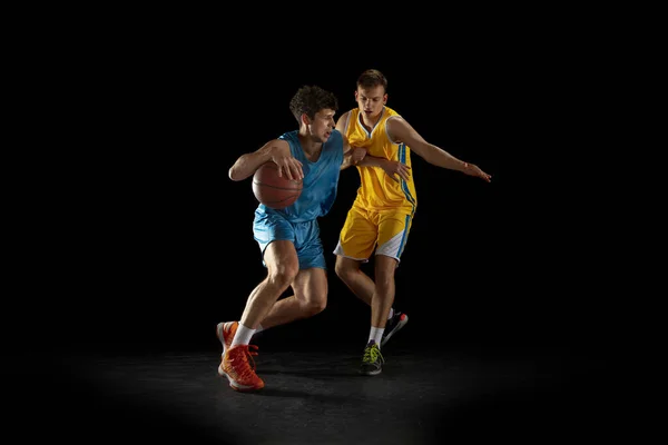 Dos jugadores de baloncesto en acción y movimiento aislados sobre fondo de estudio negro oscuro. Concepto publicitario. Fuertes atletas caucásicos practicando con pelota de baloncesto. — Foto de Stock
