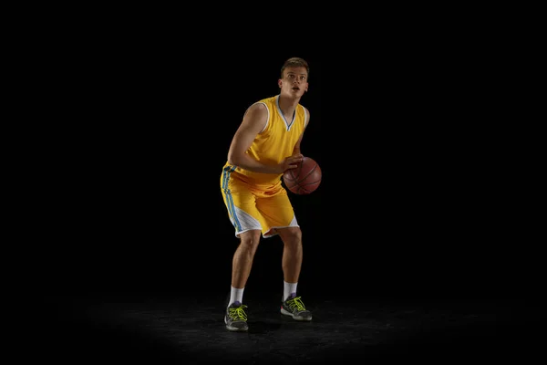 One young man, basketball player with a ball training isolated on dark black studio background. Advertising concept. Fit Caucasian athlete practicing with ball.