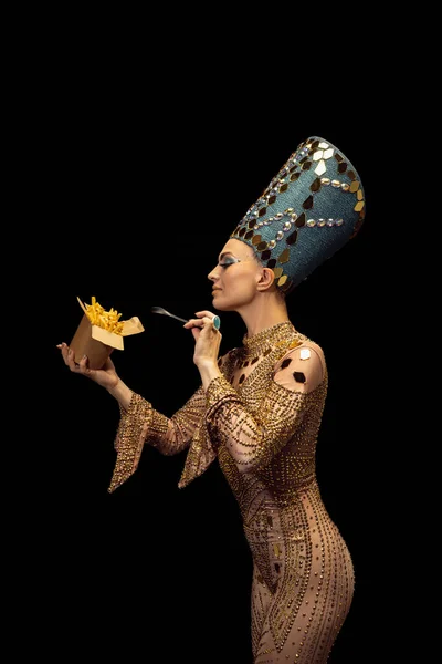 Mulher bonita nova na imagem de Nefertiti no desempenho da arte que prova o fast food moderno isolado no fundo escuro. Estilo retrô, comparação de eras, conceito de humor. — Fotografia de Stock