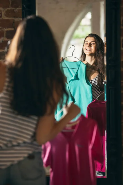 Chica joven feliz probándose nuevos trajes, preparándose para las vacaciones de verano de pie frente al espejo en casa, en el interior. Tomando la decisión correcta. Se ve emocionado, encantado. — Foto de Stock