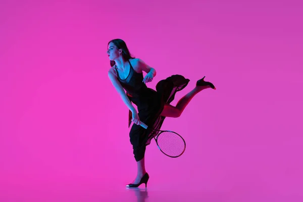 Wysokiej mody młoda kobieta w czarnej sukni wieczorowej, sukienka gra w tenisa na różowym neonowym tle. Pojęcie mody i sportu — Zdjęcie stockowe